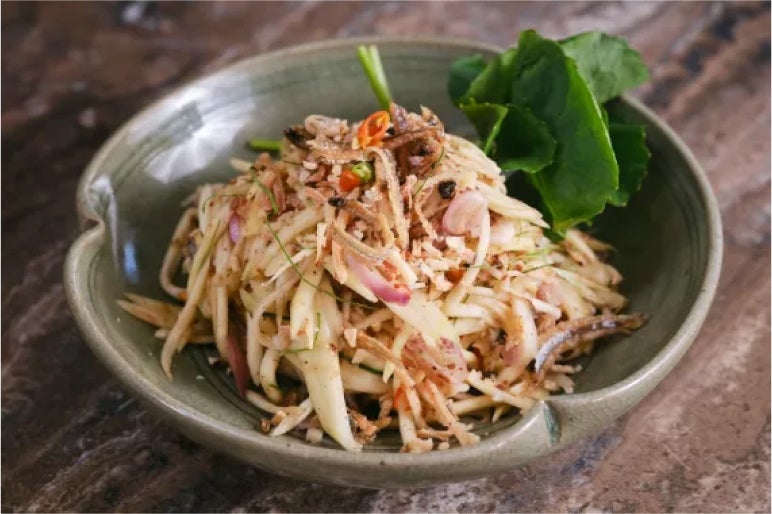 Sour mango salad with dried shrimp, red onions, and fresh herbs, garnished with leafy greens in a green ceramic bowl