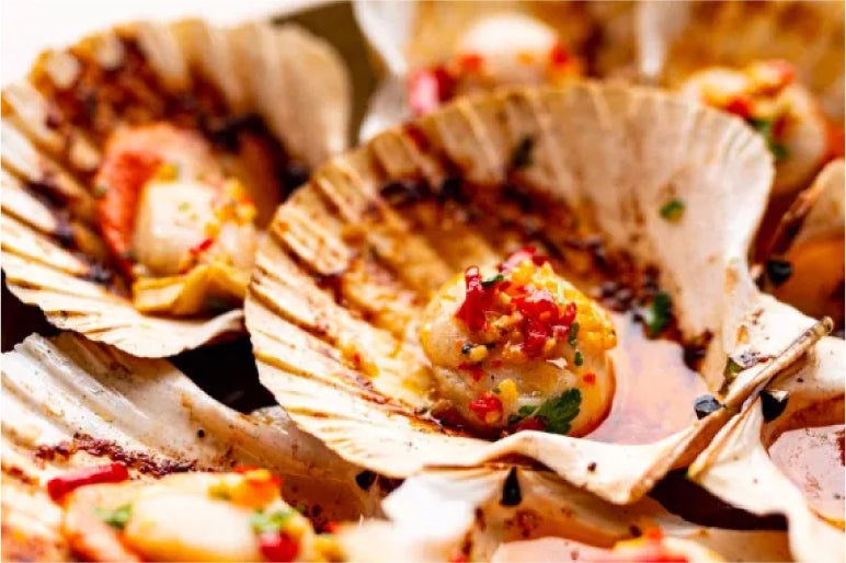 Grilled scallops in their shells, garnished with chili and fresh herbs, offering a flavorful burst of the sea and spice