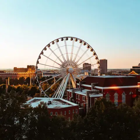 Ferris wheel at sunset with surrounding red-brick buildings and trees in the foreground