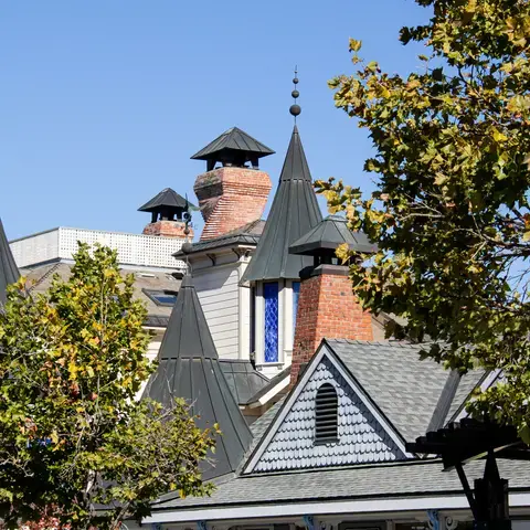 Close-up of a building with pointed turrets, brick chimneys, and decorative details, framed by leafy trees under a clear sky