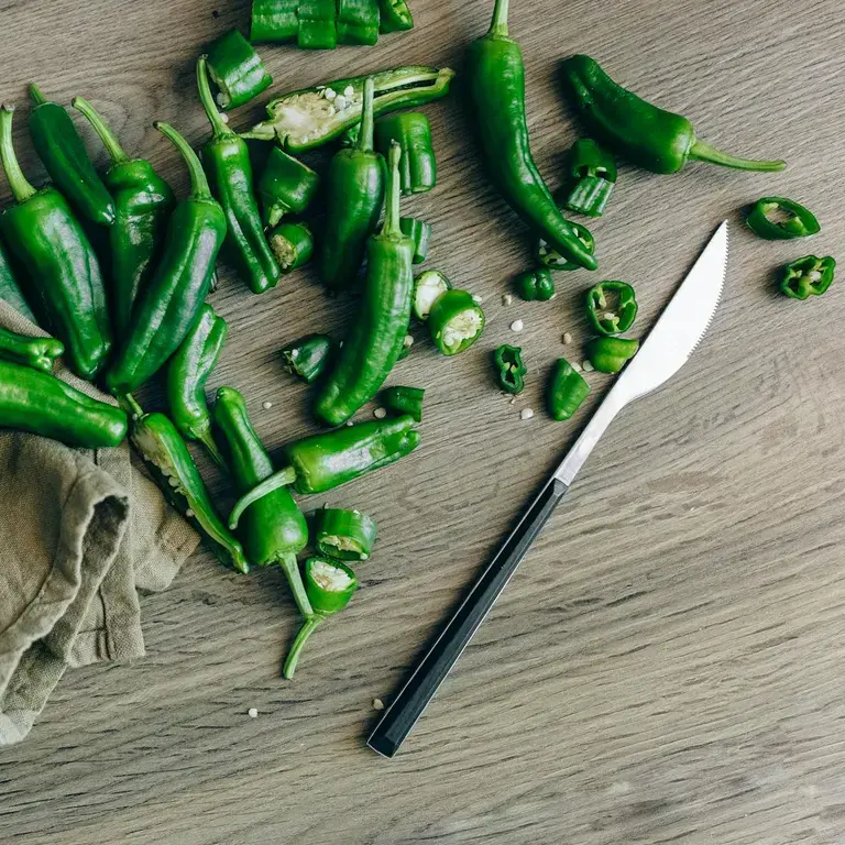 A chopping board and knife with green whole and sliced jalapeno peppers