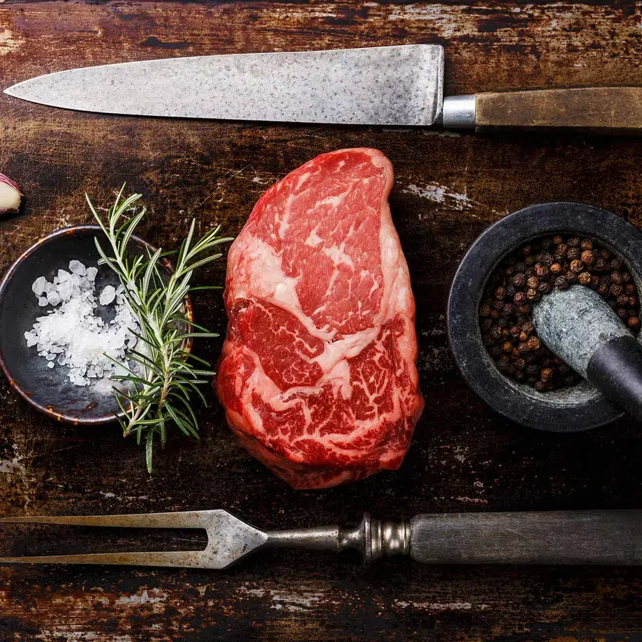 Ribeye steak on a board with seasoning, herbs, and an knife and fork.