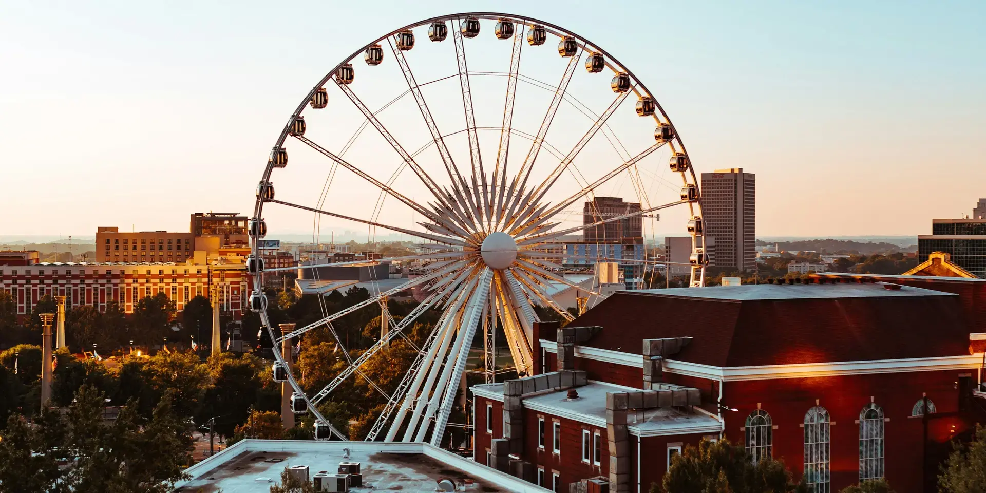 Ferris wheel at sunset with surrounding red-brick buildings and trees in the foreground