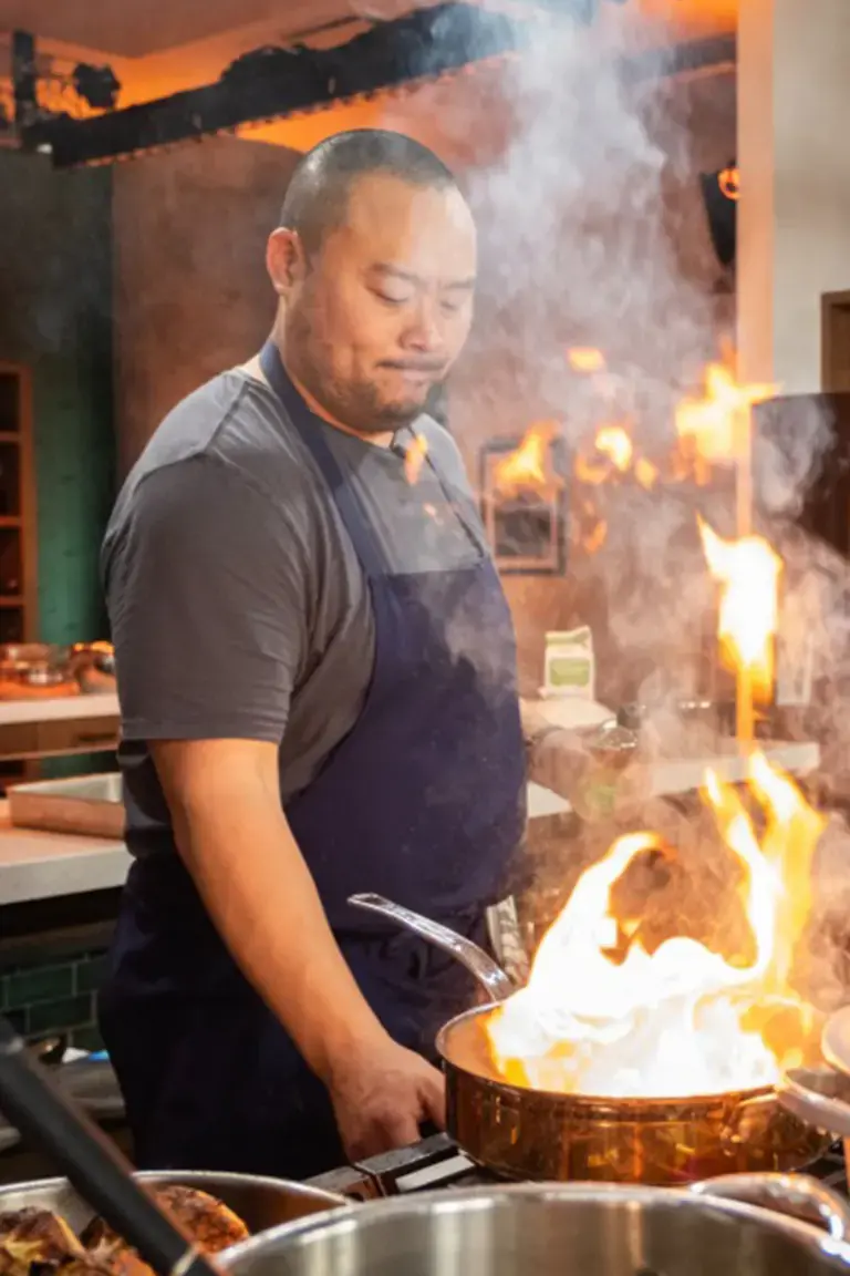 David Chang smiling in a red plaid shirt and apron pops a bottle of champagne at a festive dinner table filled with holiday