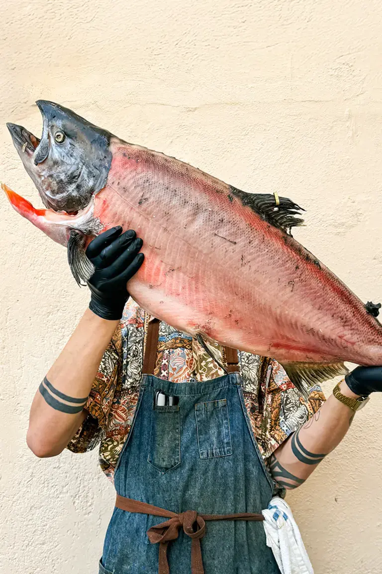 A person wearing an apron, patterned shirt, and black gloves holds a large salmon horizontally in front of their face against