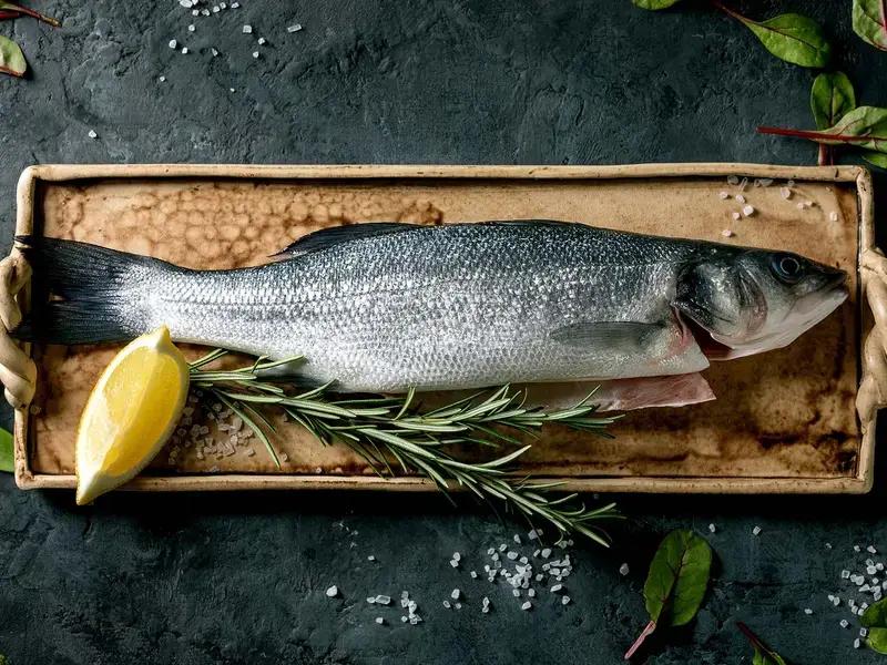 Raw fresh uncooked fish sea bass on ceramit plate ready to cook with lemon, sea salt, rosemary, green salad over dark texture